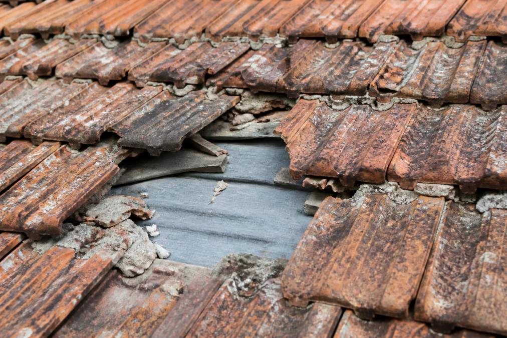 Close-up of cracked, missing, and heavily weathered clay roof tiles on an older property, showing exposed grey underlayment and deteriorated mortar joints between tiles