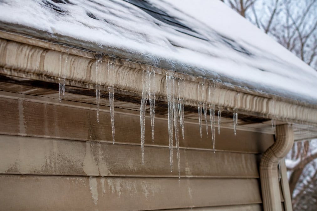 Icicles and ice dams hanging from a roof edge during winter, with visible water staining and potential leak damage on the fascia