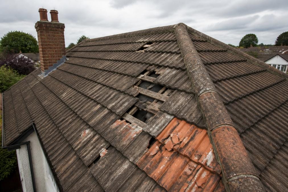Aerial view of a residential roof showing broken roof tiles, gaps between slates, and damaged clay tiles with visible deterioration against a cloudy sky