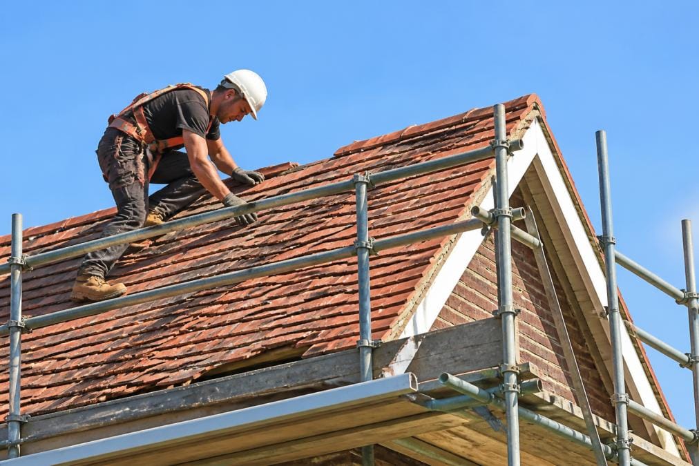 Skilled roofer working on residential pitched roof with traditional clay tiles, wearing safety harness and hard hat, working from metal scaffolding against clear blue sky