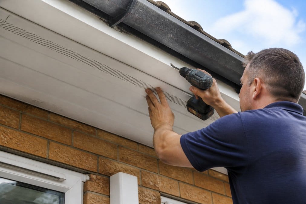 Tradesperson installing new uPVC soffit board with ventilation strips clearly visible, using proper nail spacing guidelines and applying silicone sealant at joints where boards meet