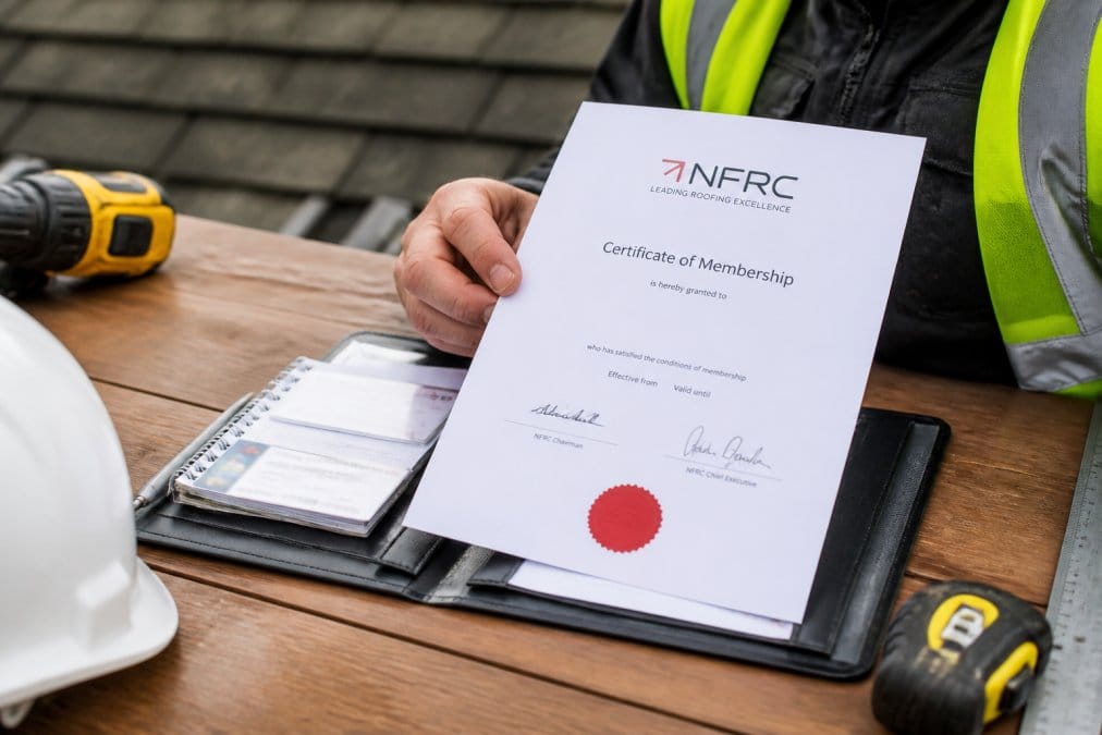 Professional roofer's hand holding NFRC certification document and credentials folder on a wooden desk with tools visible in background