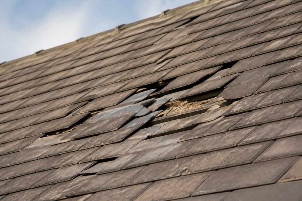 Close-up of a roof showing cracked, missing, and displaced tiles with visible damage patterns and weathering from wind exposure, photographed in natural daylight