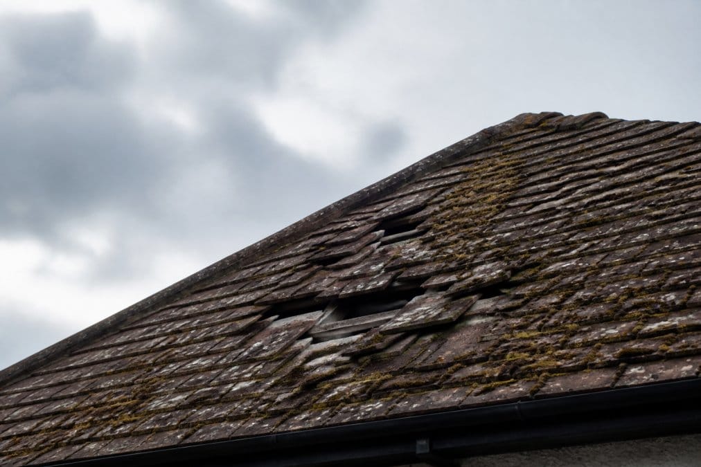 Close-up of residential roof showing visible damage with missing tiles, moss growth, weathered shingles, and overcast sky in background