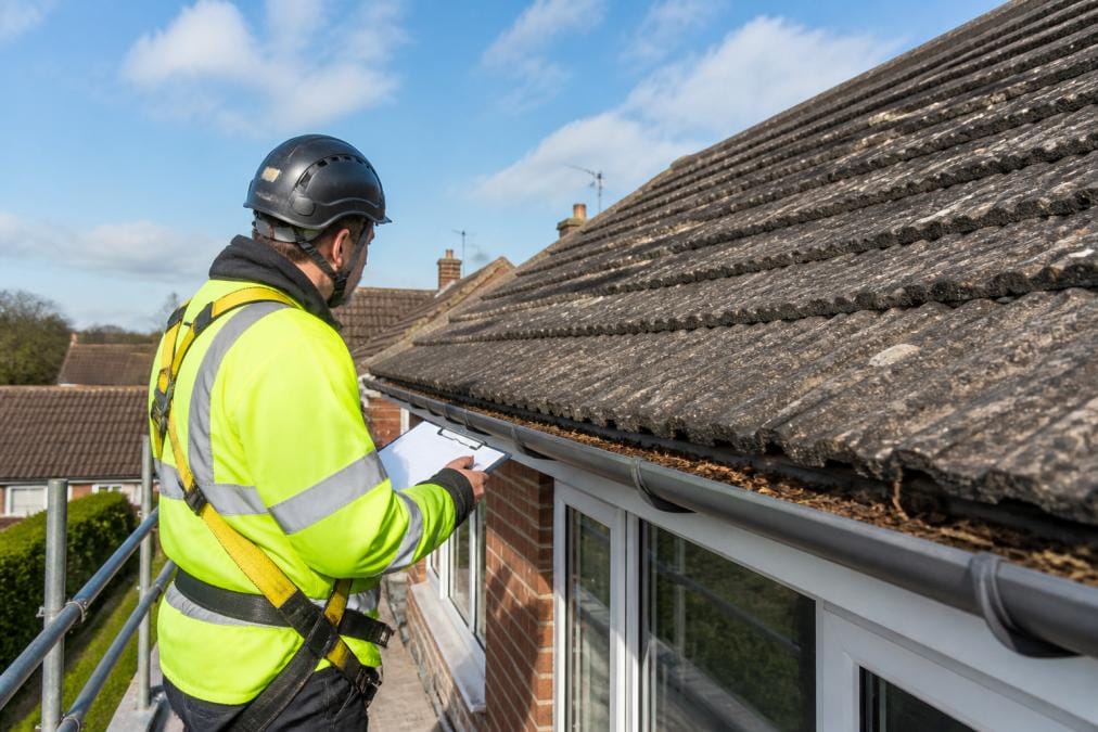 A roofer in high-visibility gear and safety harness inspecting concrete roof tiles and a gutter run on a typical British semi-detached house on a clear day, clipboard in hand