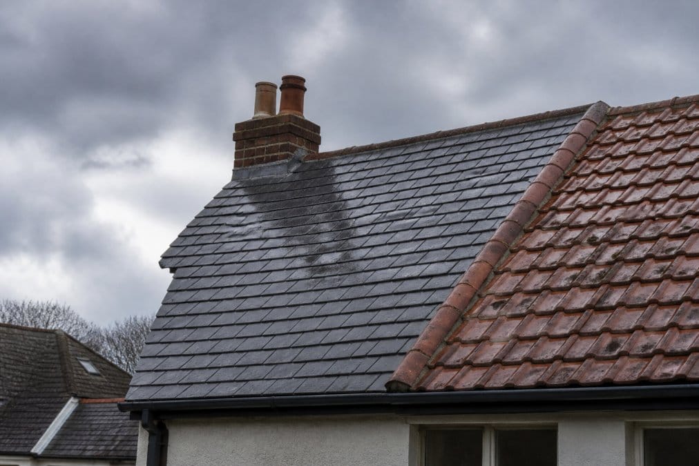 A close-up view of a UK home's pitched roof showing a mix of natural slate and clay tiles under a heavy overcast sky, with damp patches on the tiles indicating recent rain