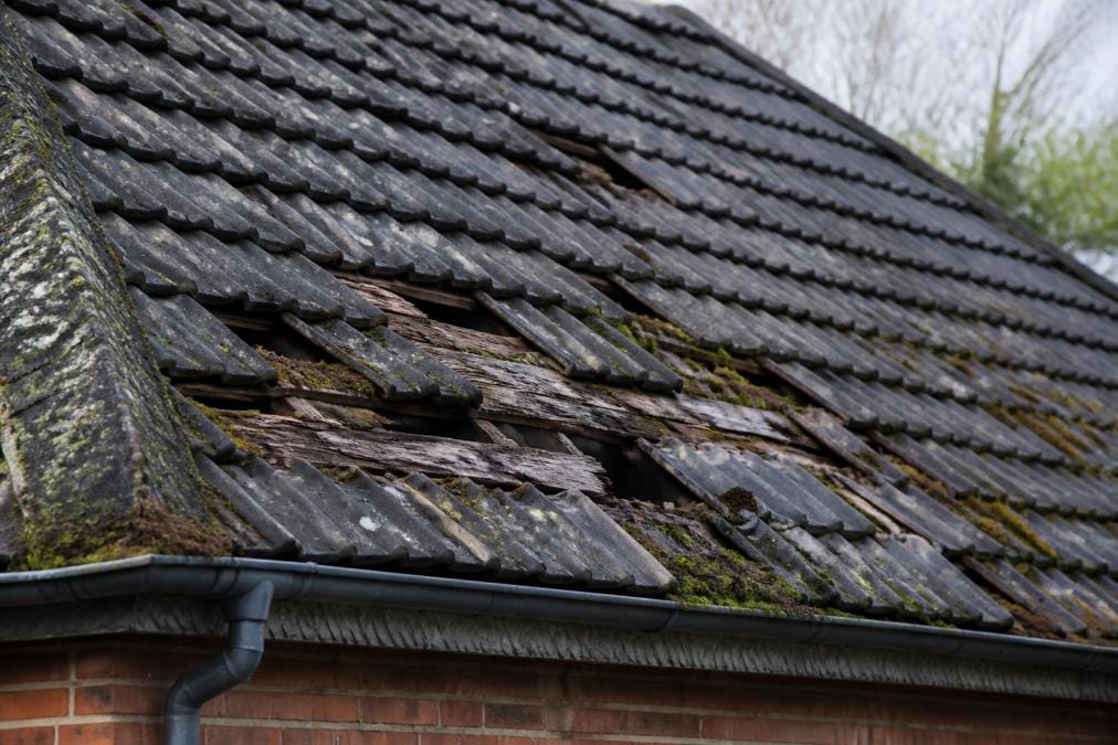 Close-up of a roof showing visible damage including missing tiles, cracked shingles, moss growth, and weathered surfaces with clear structural wear