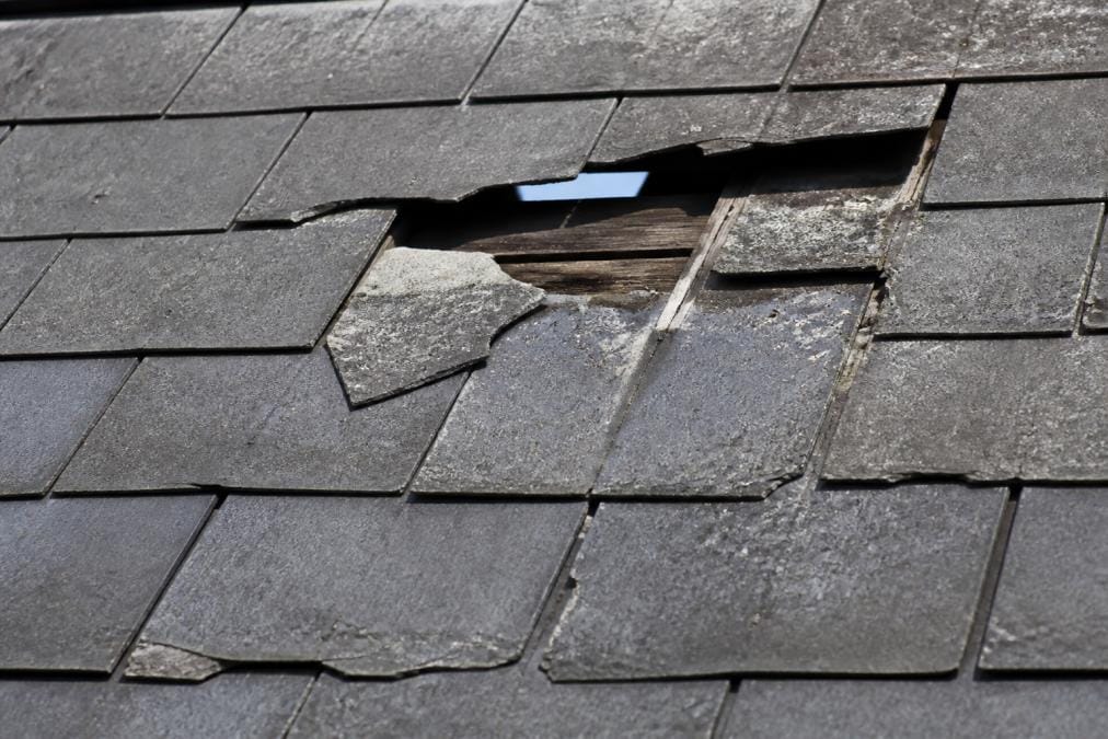 Close-up of roof tiles showing cracked shingles, missing sections, and visible deterioration with gaps allowing daylight through