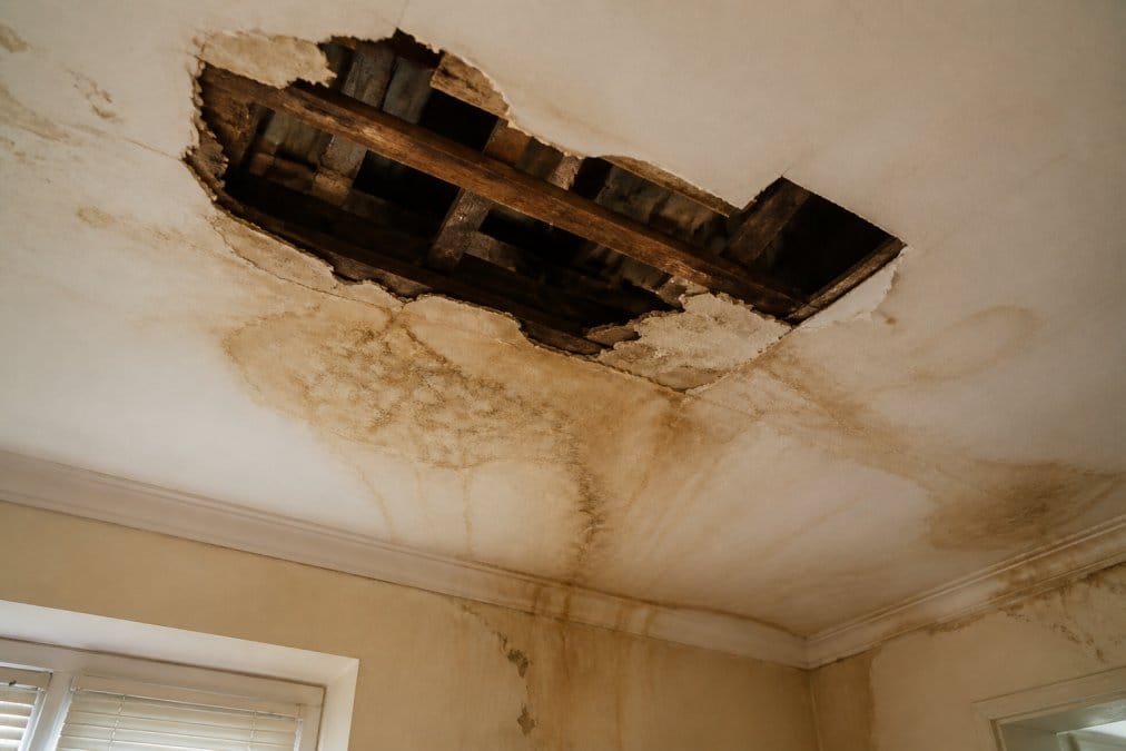 Close-up photograph of a damaged UK residential roof with missing tiles and brown water staining spreading across the ceiling below, shot in flat daylight