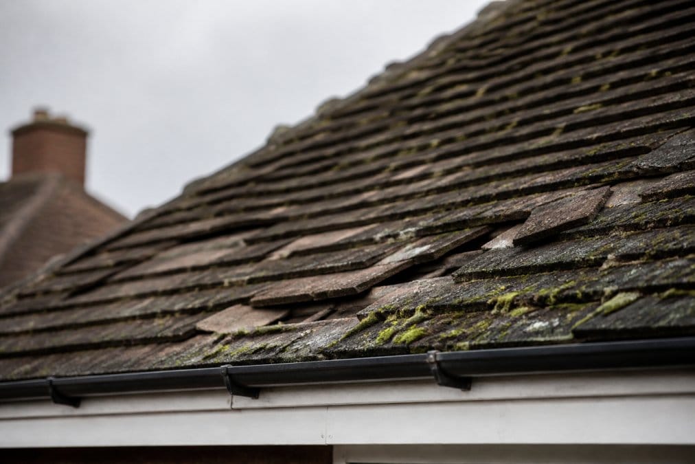Close-up photograph of an ageing UK residential roof with missing, cracked and curling tiles, moss growth between them, shot from a low angle on an overcast day
