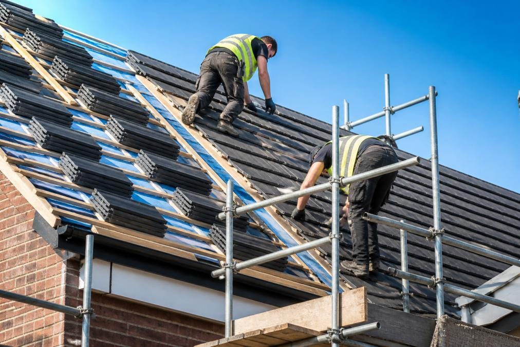 A roofing team in safety gear installing new tiles on a residential house roof, with scaffolding visible and a clear blue sky behind them