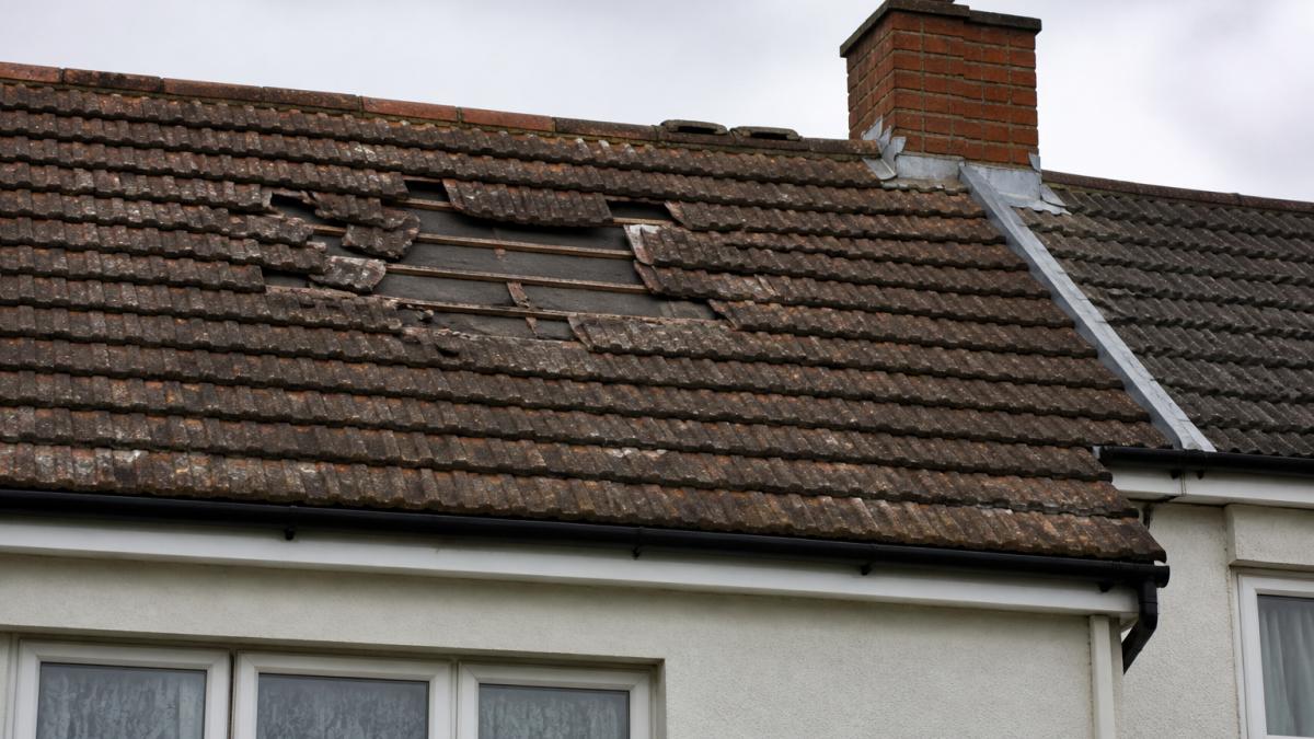Close-up of a weathered semi-detached house roof with several slipped and cracked clay tiles, showing the exposed underlay beneath, taken on an overcast day