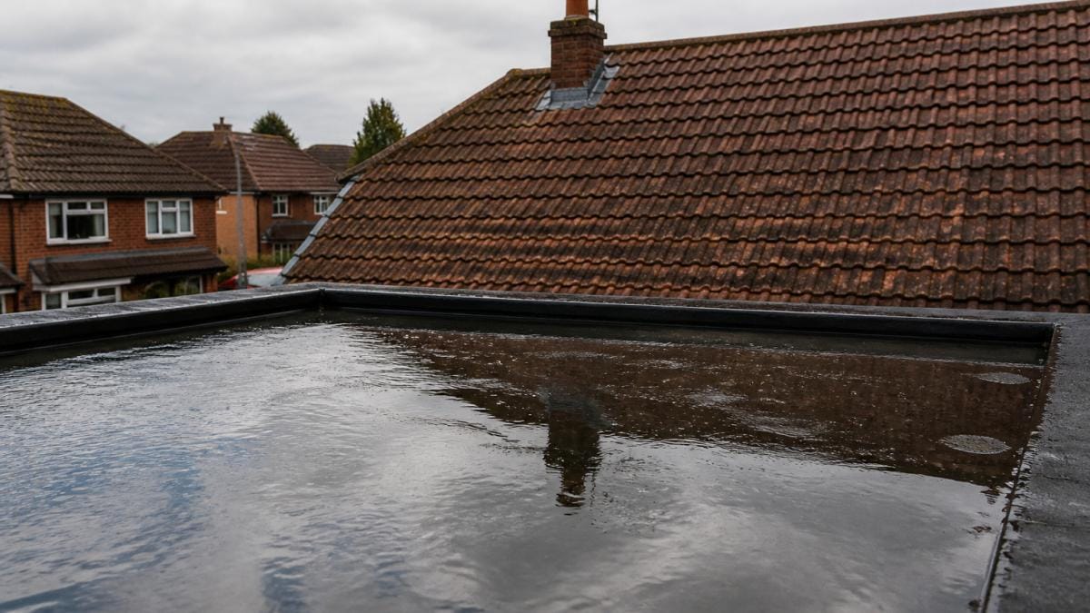 Close-up photograph of a flat roof holding shallow rainwater with a pitched tiled roof visible behind it, overcast daylight, suburban UK housing