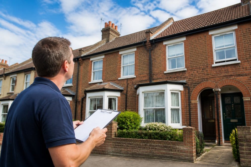 A homeowner standing on a driveway looking up at a terraced house roof, clipboard in hand, checking gutters and tiles on a clear day